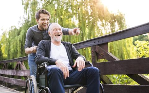 Junger Mann schiebt Rollstuhl mit älterem Herrn über eine Brücke in einem Park